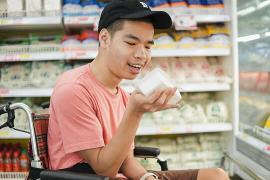Happy young Asian man in a wheelchair reading a nutrition label on a food pack at the supermarket, concept of accessibility, healthy living, and inclusion.