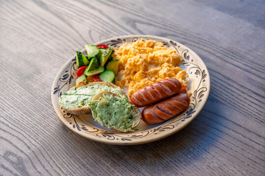 A hearty breakfast plate with scrambled eggs, grilled sausages, fresh cucumber and tomato salad, and toasted bread with herb butter