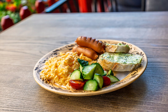 A hearty breakfast plate with scrambled eggs, grilled sausages, fresh cucumber and tomato salad, and bread with herb butter on a wooden table