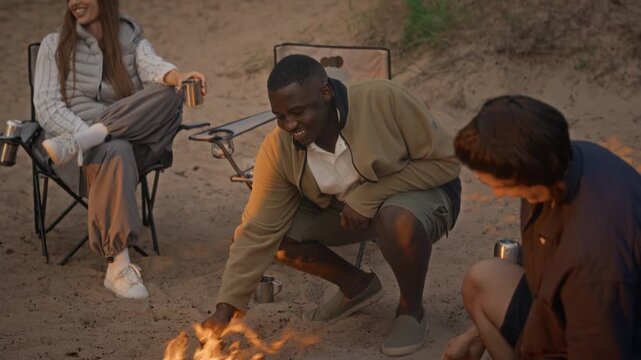 Diverse young adults roasting marshmallows over campfire on sandy beach while camping. Clip is perfect for outdoor recreation, travel gear, and youth lifestyle marketing campaigns