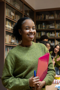 High school student smiling, holding a file and pen, with classmates in the background