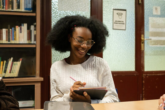 Young woman with afro hair and glasses writing on tablet, laughing during study