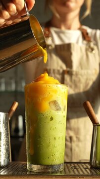 Female barista pours vibrant mango puree over a green iced drink in a glass, showcasing a colorful beverage creation process in a modern cafe setting