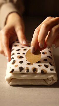 Hands of a female wrapping a gift with spotted wrapping paper, placing a decorative circle on top, showcasing a cozy indoor setting with soft lighting and neutral tones