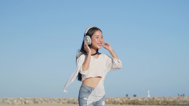 Happy woman on summer beach loves to listen to music and dance with headphones, cheerful female enjoying outdoor leisure on sunny day, beautiful Asian person having fun during vacation.