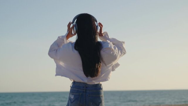 Cheerful attractive beautiful woman on tropical beach likes to listen to music on vacation holiday, dancing with raised hands wearing wireless headphones near ocean during summer sunset.