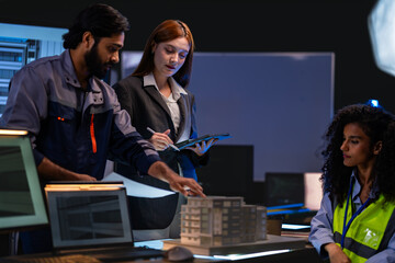 Construction team members review a building scale model and blueprints in a dark operations center...