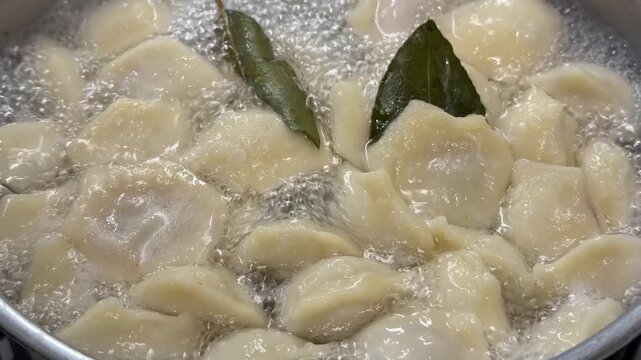 Fresh homemade dumplings boiling in a pot of water, bay leaves floating on surface, steaming hot traditional meal, close up view of cooking process