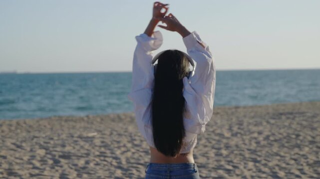 Happy young woman enjoying music and dancing on sunny beach while wearing headphones and white shirt, female model with long dark hair looking at calm blue ocean during summer holiday.
