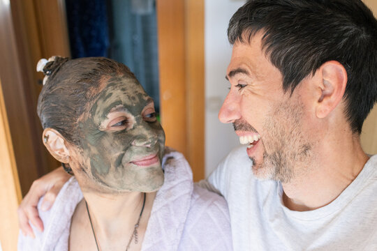Couple laughing together, a woman wearing a green clay beauty mask on her face, sharing wellness and self-care