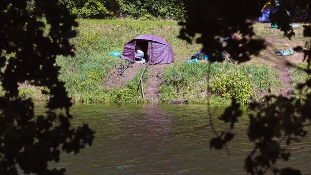 Fisherman camping in a camouflage tent on a grassy riverbank on a sunny summer day
