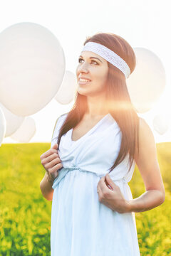 A happy young woman in a white dress and headband holds white balloons in a sun-drenched green field, looking up with a joyful expression. The warm sunset creates a golden glow.