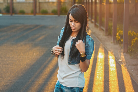 Stylish young woman in denim vest and white shirt, looking down, posing on an outdoor sports court during warm golden hour. Casual street style, long dark hair.