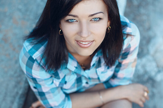 High-angle top view portrait of a beautiful young woman with dark hair and striking blue eyes looking up and smiling warmly at the camera, wearing a casual blue plaid shirt outdoors.