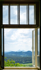 Fototapeta premium Scenic landscape view of Bezdez castle twin peaks framed by an open old wooden window looking out over green forests and hills under a cloudy summer sky.