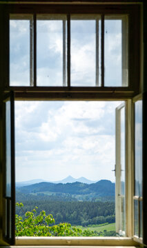 Scenic landscape view of Bezdez castle twin peaks framed by an open old wooden window looking out over green forests and hills under a cloudy summer sky.