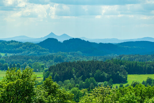 Scenic landscape view of rolling green hills, dense forests and meadows with the iconic twin peaks of Bezdez castle silhouetted on the distant horizon under a cloudy summer sky.