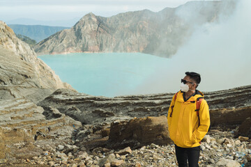 A masked Asian hiker in a yellow jacket and orange backpack stands on rocky ground overlooking a turquoise volcanic Ijen crater lake, surrounded by misty cliffs and dramatic, remote mountain scenery e © Alfian Muzaki