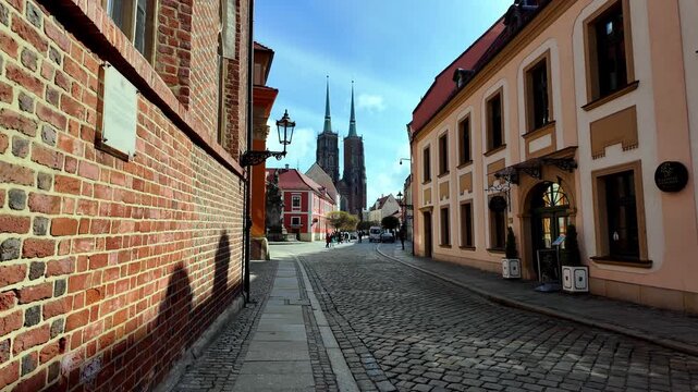 Video footage of a cobblestone street in Ostrow Tumski, Wroclaw, featuring a red brick wall on the left, historical townhouses, and the towers of Saint John the Baptist Cathedral in the background in 