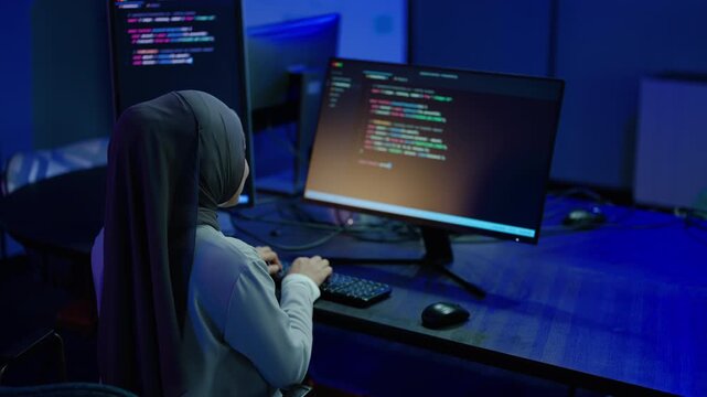 Dedicated Muslim female programmer wearing a hijab coding software on dual computer monitors in a dark modern office at night. IT developer typing data. Cyber security professional working late.