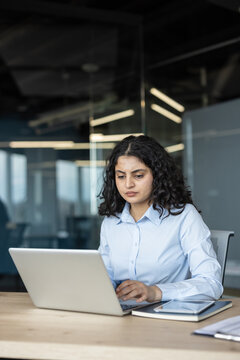 Young businesswoman with dark curly hair sitting at a wooden desk, concentrating on her work while typing on a laptop in a modern corporate office environment