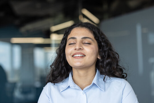 Young businesswoman with eyes closed and a gentle smile practices mindful deep breathing at her desk, finding calm, focus, and stress relief in a modern office setting