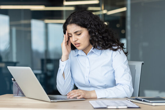Young businesswoman working on laptop experiencing severe headache and migraine, touching her temple while feeling exhausted and overwhelmed with work in a modern office