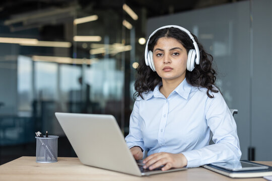 Young businesswoman wearing white headphones, focusing on a laptop, and working professionally at her desk in a modern corporate office, embodying remote work and technology
