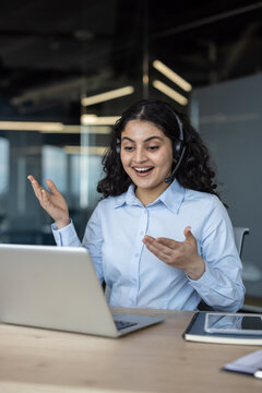 Young indian woman with headset smiling and gesturing during a video call, working on a laptop in a modern office setting, providing online support and consulting remotely