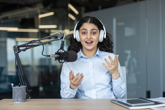 Young woman wearing headphones and speaking into a professional microphone, actively creating audio content for a podcast or radio broadcast in a contemporary office setting