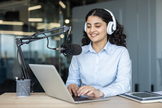 Young smiling woman wearing headphones, actively engaging in a podcast recording session, using a professional microphone and laptop in a modern studio setting