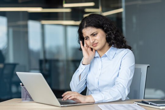 Young business professional experiencing intense headache or migraine while working on a laptop at her office desk, expressing discomfort and exhaustion