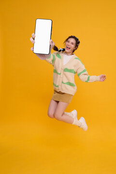 Young woman jumping while showing smartphone blank screen expressing mobile marketing digital promotion social media engagement online communication app advertising and energetic lifestyle.