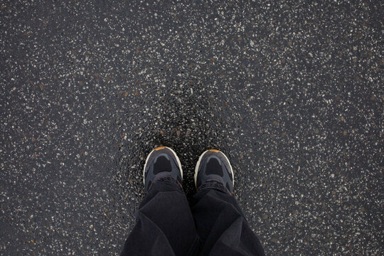 Feet of woman in sneakers and loose grey jeans standing on wet asphalt after the rain close up top view. Human in the city, walk in rainy weather, minimalistic background