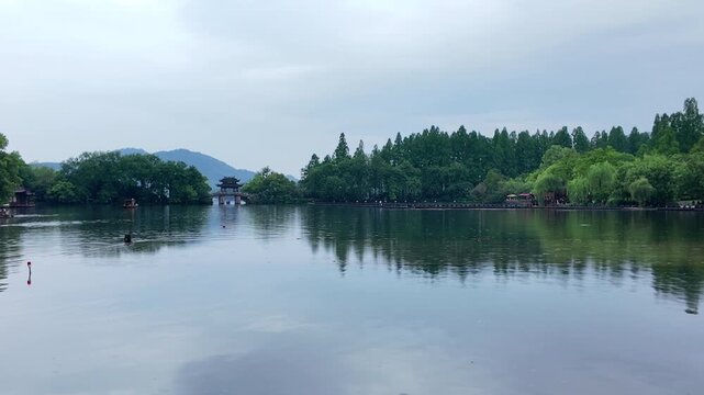 A serene lake landscape with dense green trees, distant mountains, and a traditional pavilion under an overcast sky.