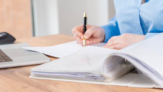 Person writes with a pen in a blue shirt. Laptop rests on a wooden desk. Open notebook lies beside their work. They concentrate fully on the task. This scene suits office or academic settings