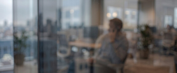 Fototapeta premium A blurred image of a person seated in an office, with modern workspace elements in the background and city views visible through the windows.