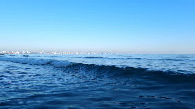 A serene view of the ocean with gentle waves approaching the shore, featuring a distant city skyline.