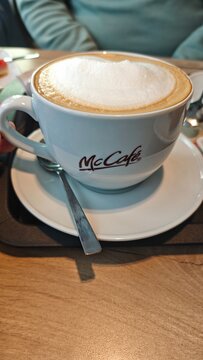 Berlin, Germany - December 01, 2025: McCafe cappuccino served in a white ceramic cup on a round plate with a stainless steel spoon on a wooden table