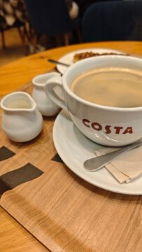 Gdansk, Poland - July 18, 2026: Coffee cup with Costa logo, milk jug, and dessert plate on wooden table in a cafe setting, spoon and napkin included
