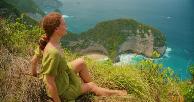 Young woman sits on a grassy cliff edge admiring the view of Kelingking beach Nusa Penida