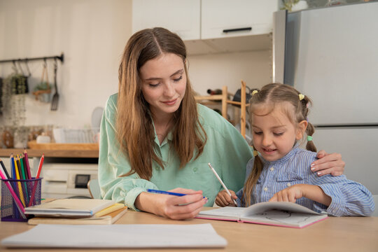 Mother embracing her little daughter while helping her with school homework at home. Loving mother sitting at kitchen table supporting cute daughter with homeschooling assignment and reading notebook.