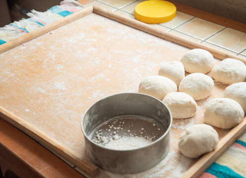 Raw dough balls on a floured wooden board beside a metal sifter and rolling pin, rustic kitchen scene suggesting homemade bread or pastry preparation and baking process
