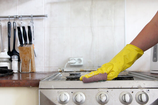 Person wearing a yellow rubber glove and using a sponge to clean a dirty gas stove in a home kitchen, representing household chores, hygiene, and spring cleaning.