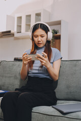 Beautiful young asian woman sitting on sofa and holding joystick playing online video game in living room at house © ARMMY PICCA