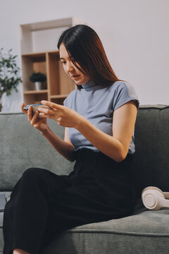 Freelance Asian woman holding mobile phone typing on laptop and working from home. Happy young girl sitting on sofa in living room.