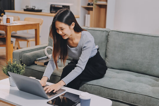 Freelance Asian woman holding mobile phone typing on laptop and working from home. Happy young girl sitting on sofa in living room.