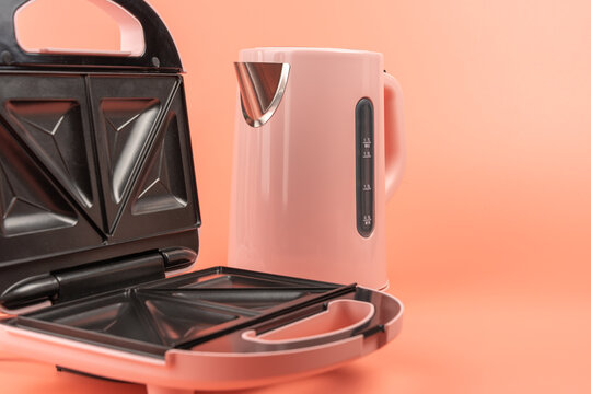 An open pink sandwich maker with black triangle plates sits beside a pink electric kettle on a solid coral background, closely framed.