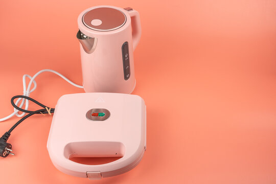 A pink electric kettle stands beside a closed pink sandwich maker on a solid coral background, both connected by their power cords.