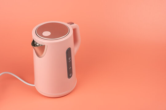 A closer three-quarter view of a pink electric kettle shows its brushed steel lid and spout against a solid coral background.